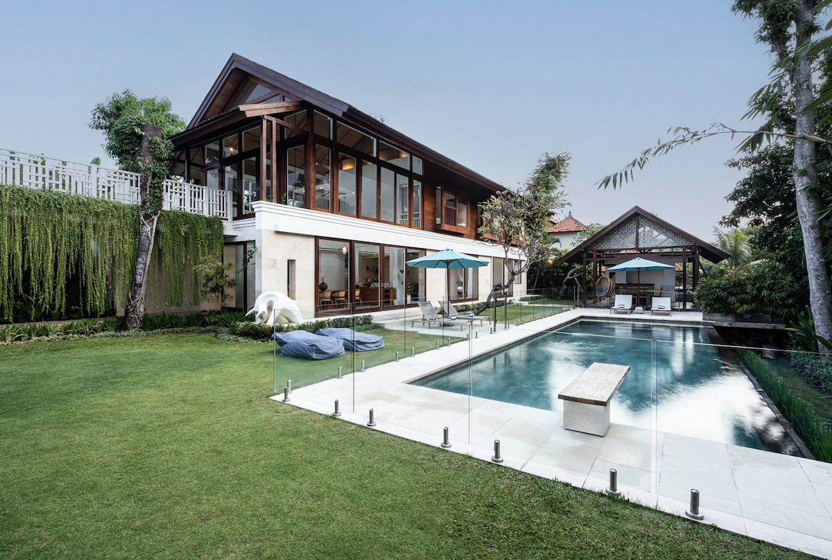The spacious villa features a modern architectural design with large glass windows, surrounded by lush greenery. An infinity pool reflects tranquil water, complemented by a wooden deck and lounge chairs. The outdoor area offers an inviting atmosphere for relaxation and enjoyment.