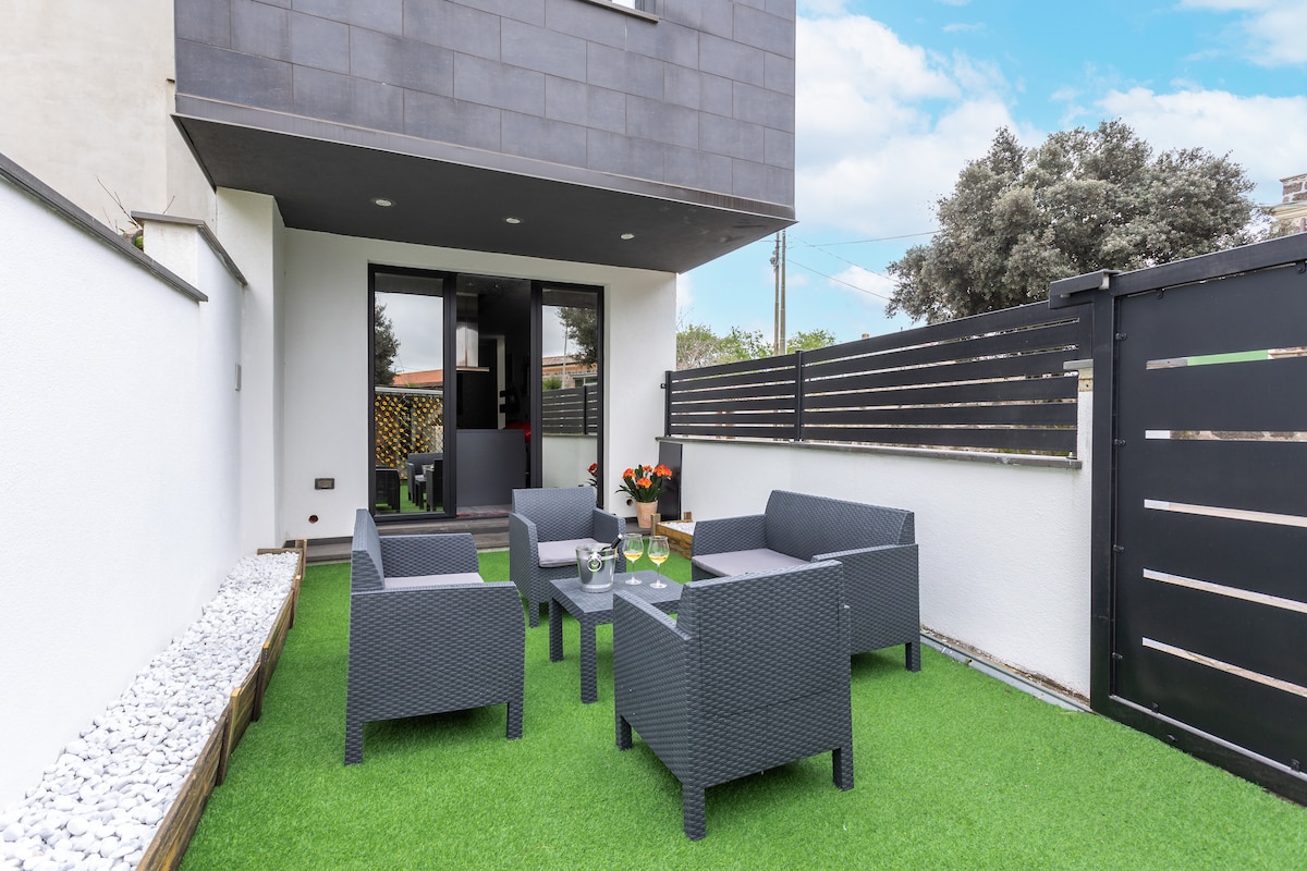 An outdoor seating area features comfortable grey wicker furniture arranged on manicured green turf. Potted flowers add a splash of color to the space, which is enclosed by modern fencing and a clean white wall, creating a private retreat right outside the entrance.