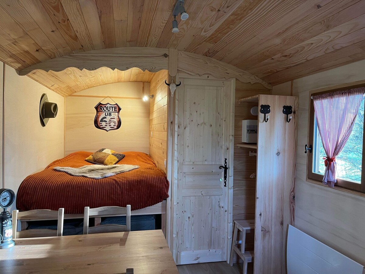 The interior of a rustic cabin features a cozy sleeping area with a spacious bed adorned in warm, earthy tones. A wooden table and chairs are visible, alongside a small kitchenette. Natural light filters in through a nearby window, illuminating the wooden paneling of the walls.