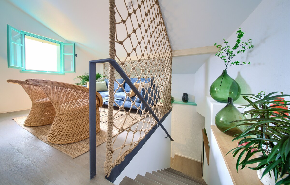 A staircase leads to a modern living space with light streaming through open turquoise windows. Decorative elements include a woven seating area and green glass vases, complemented by plants adding a touch of freshness.