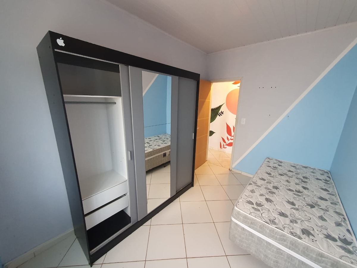 A bedroom is presented featuring a spacious bed dressed in patterned bedding. A large mirrored wardrobe with a sleek design stands against the wall. Light blue paint accents add a calming touch to the room, illuminated by natural light from a nearby door.