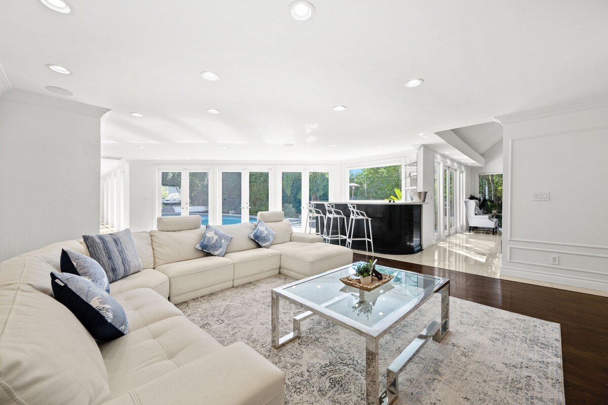 A spacious living area features a large, light-colored sectional sofa with decorative pillows arranged for comfort. A sleek glass coffee table sits atop a soft area rug, while abundant natural light pours in through multiple sliding glass doors leading to the outdoor space.