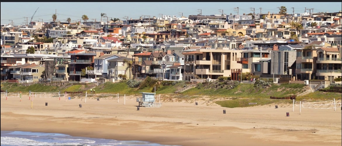 A view of beachfront properties extends along the shoreline, showcasing a variety of architectural styles. The sandy beach is visible in the foreground, with rows of palm trees lining the path. The cityscape features a mix of two to three-story homes with numerous balconies and terraces.