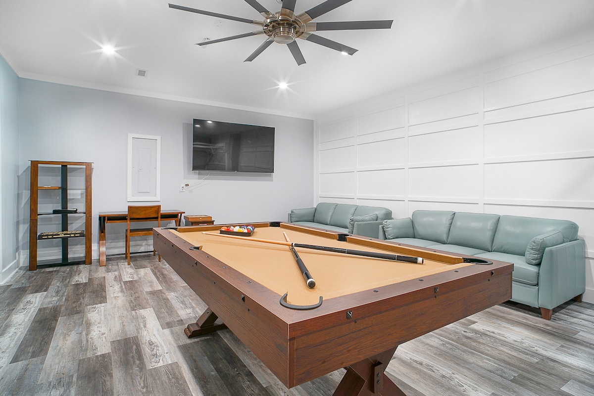 A spacious game room is highlighted by a billiards table at its center, complemented by a modern ceiling fan. Comfortable seating is provided with light green couches, while a flat-screen TV is mounted on the wall, and a small desk is positioned in the background.