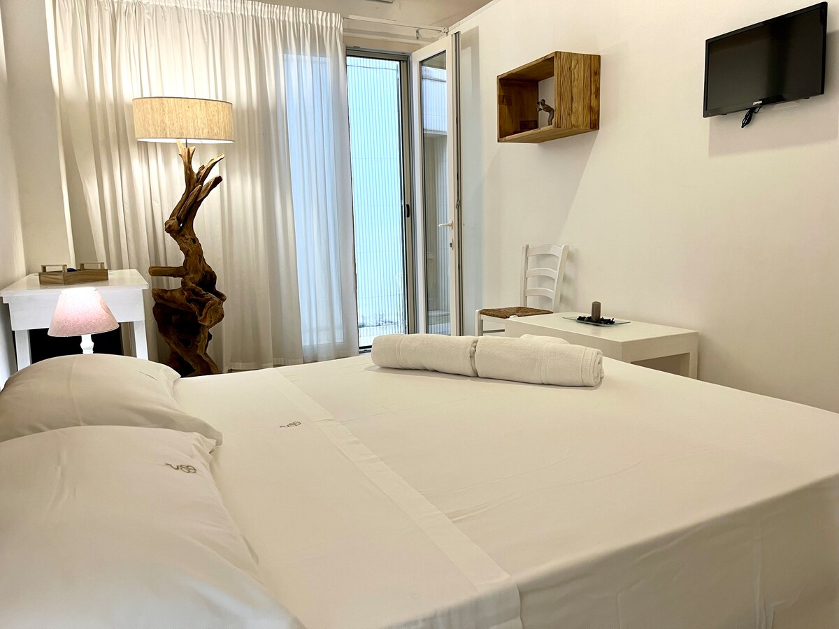 A spacious sleeping area features a large bed with crisp linens. Natural light is welcomed through glass sliding doors, leading to a small outdoor area, while a wooden lamp and TV are positioned thoughtfully within the room.