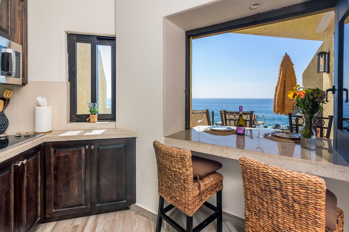 A bright kitchen area features a window that opens to stunning ocean views. Barstools with woven seats provide seating at the granite counter, where a table is set for dining. Natural light fills the space, enhancing the connection to the seaside scenery.