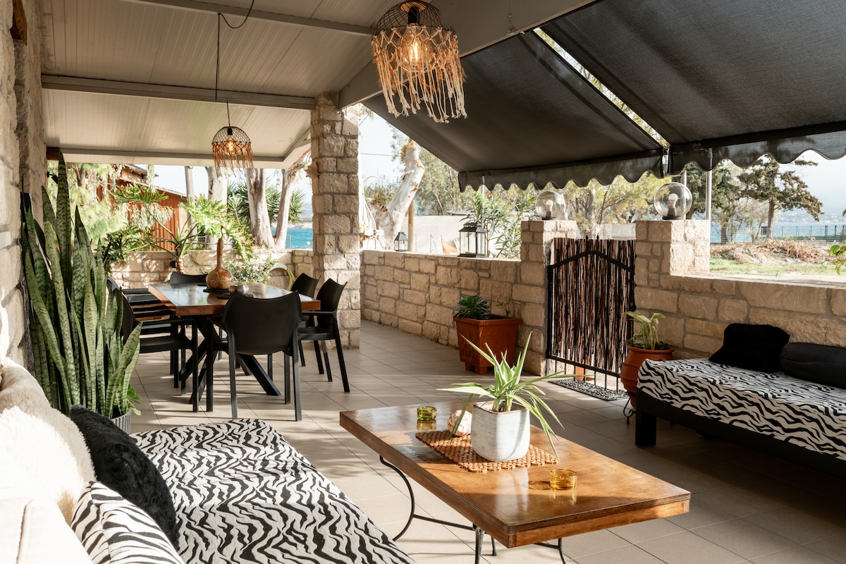 A spacious outdoor patio features a large dining table surrounded by black chairs. Soft seating options include patterned loungers. Green plants and decorative elements add a natural touch, while overhead lighting complements the shaded area, creating a comfortable gathering space with views of the surroundings.