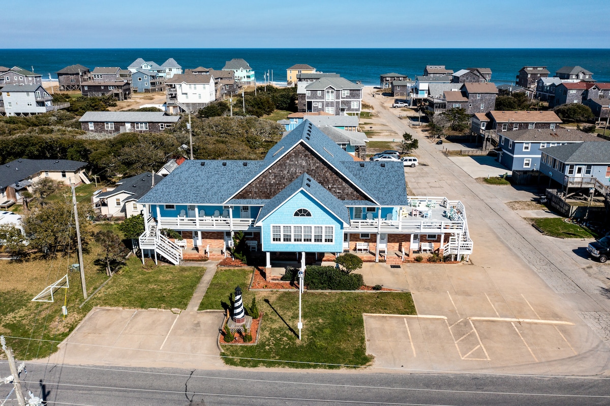 An aerial view captures a blue condotel with white railings, positioned near the coastline. Surrounding buildings are visible, illustrating a coastal community feel. The ocean glistens in the background, while a large lawn area is present in front of the property, offering space for outdoor activities.