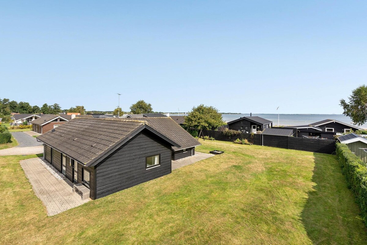 An overview of the property is presented, showcasing a spacious lawn bordered by trees. The house is depicted with large windows, and a paved area is visible. The distant view of the water can be observed, illustrating the proximity to the beach.