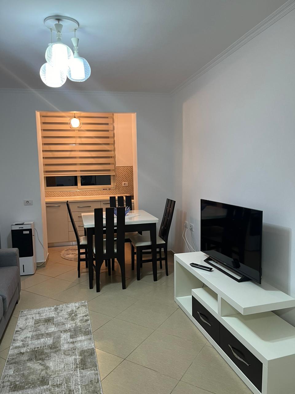 A cozy dining area features a sleek table surrounded by four black chairs. A modern television is positioned on a low white unit, and an area rug lies on the tiled floor. Soft lighting is provided by three pendant lamps overhead, adding warmth to the space.