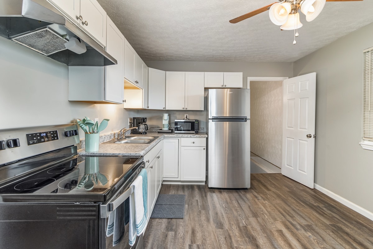 A fully equipped kitchen features modern appliances including a stainless steel refrigerator and oven. Grey cabinetry complements the light walls, while a ceiling fan enhances ventilation. A small rug is positioned on the floor, and a doorway leads to another room.