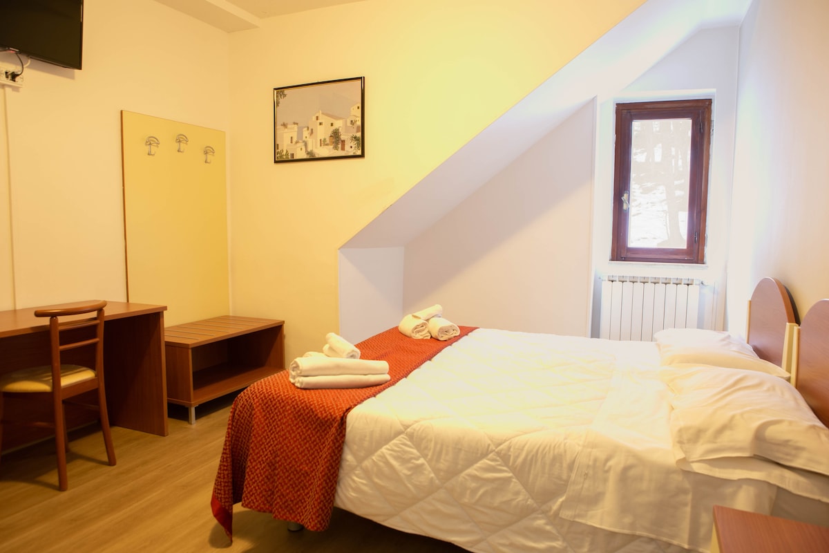 A cozy guest room features a spacious bed dressed in crisp white linens and complemented by a warm orange blanket. A wooden desk and chair provide a workspace, while a window allows natural light to fill the room. Wall hooks are available for convenience.
