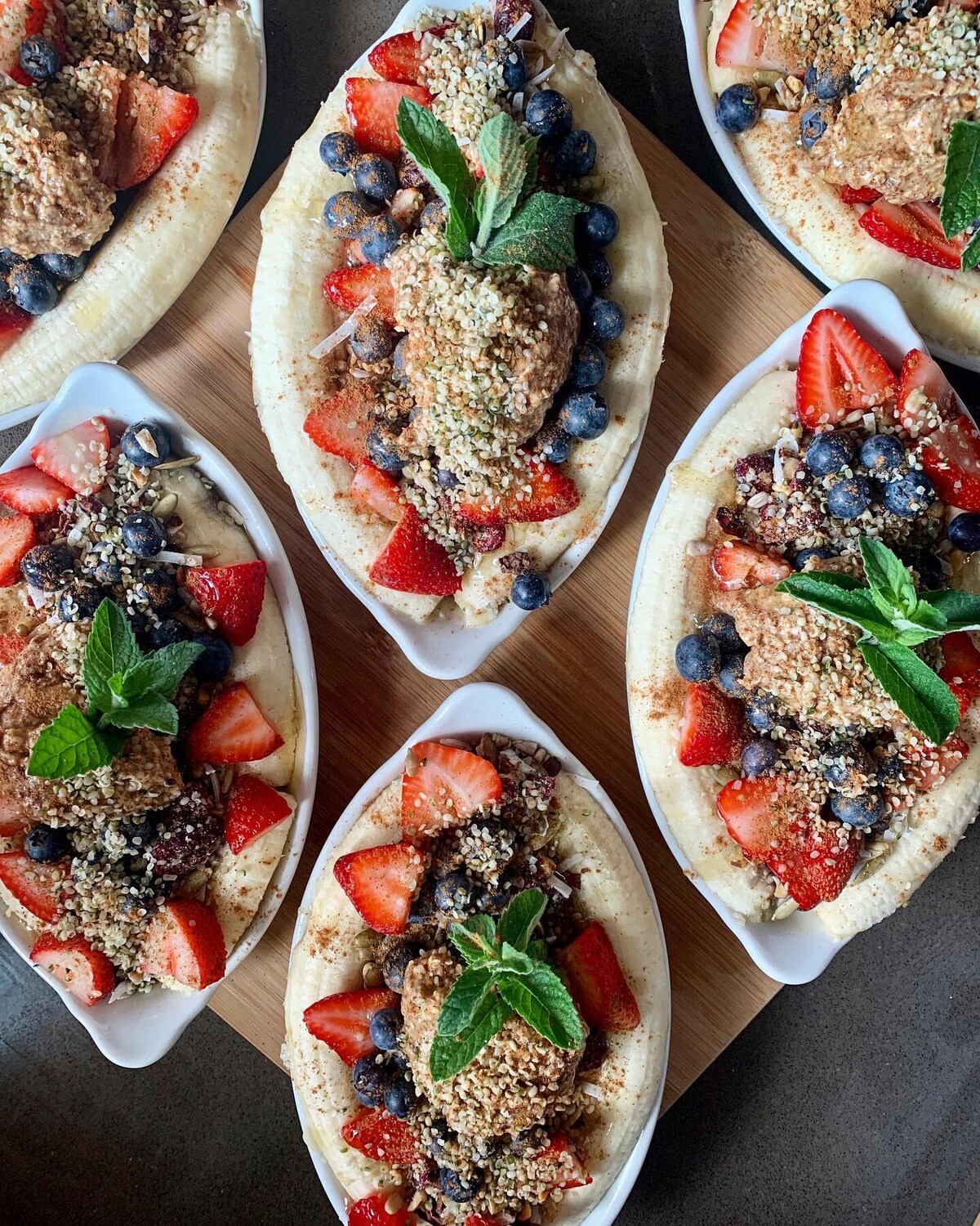 Multiple servings of banana boat desserts are arranged on a wooden tray. Each dish features sliced bananas topped with blueberries, strawberries, a dollop of nut butter, and sprinkled with granola. Fresh mint leaves are placed on top, enhancing the presentation.