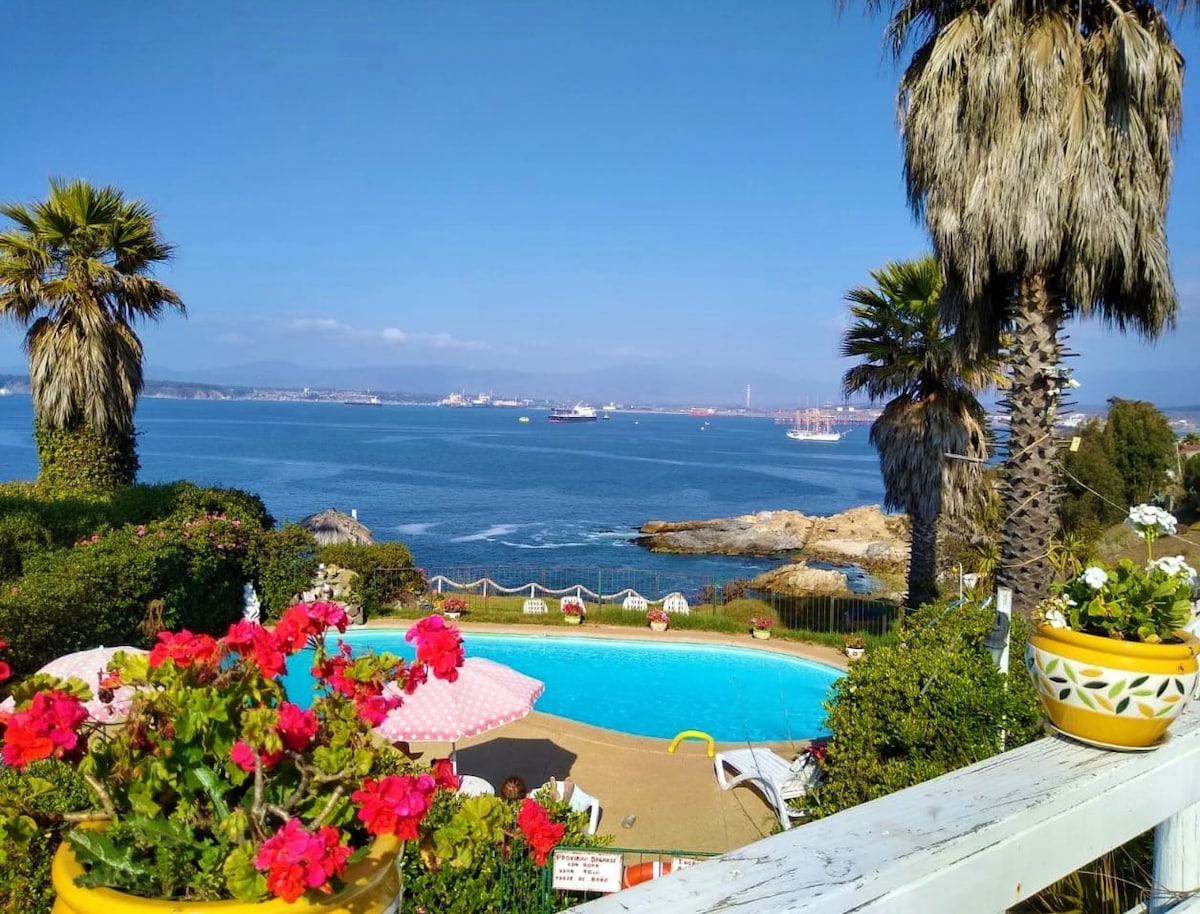 A tranquil pool area is surrounded by vibrant flowers and lush palm trees, overlooking a serene bay. The clear blue water reflects the sunny sky, while boats can be seen on the horizon, adding to the coastal ambiance.