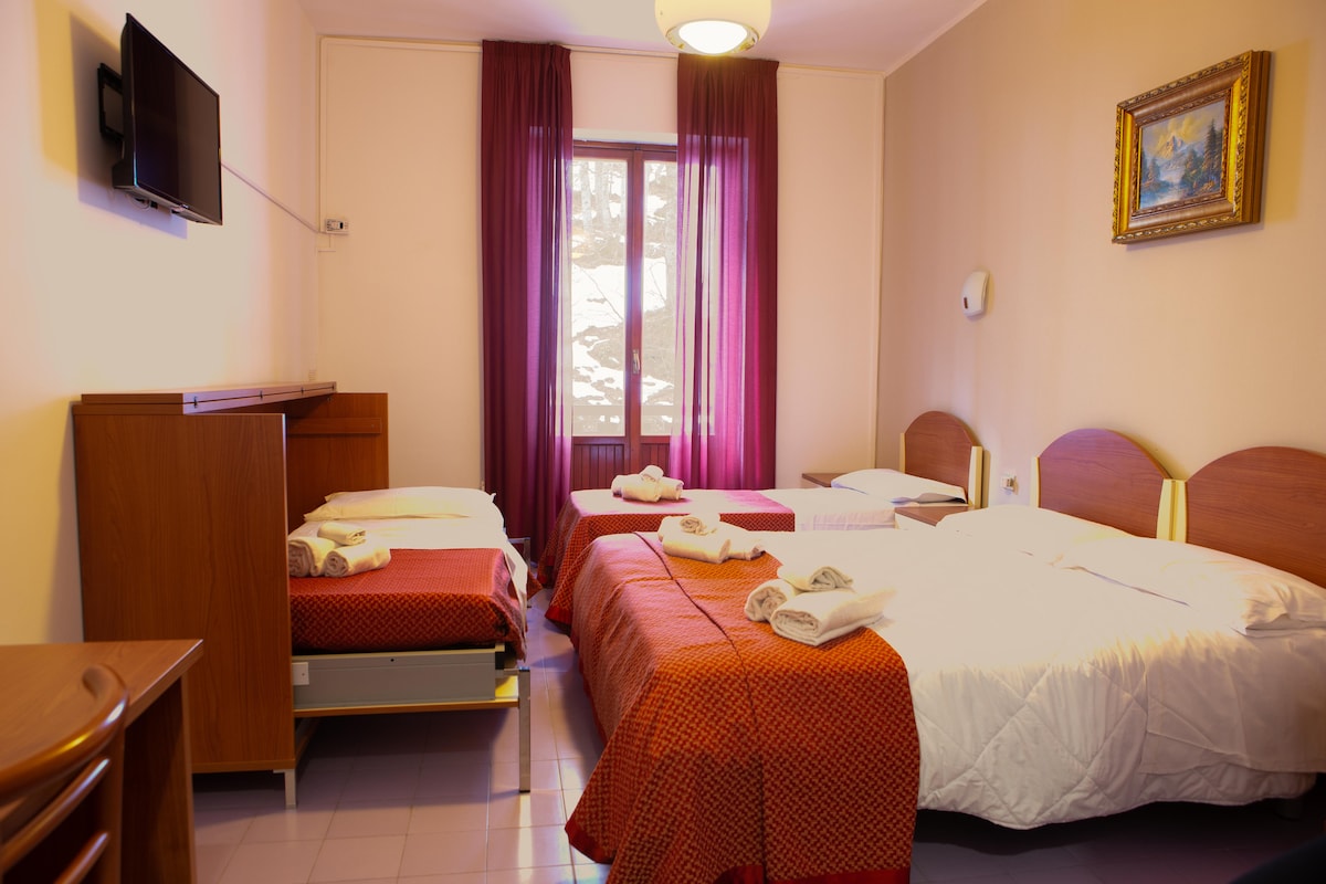 A brightly lit guest room features three beds, including two with distinct white linens and a third with a red cover. A wooden dresser and TV are visible, with a large window dressed in purple curtains allowing natural light to enter the space.