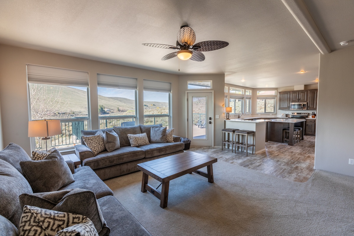 A spacious living area features large windows allowing natural light to fill the room. Comfortable seating is arranged around a wooden coffee table. The kitchen is visible in the background, showcasing modern cabinetry and an island with seating. Expansive views of the surrounding landscape are available.