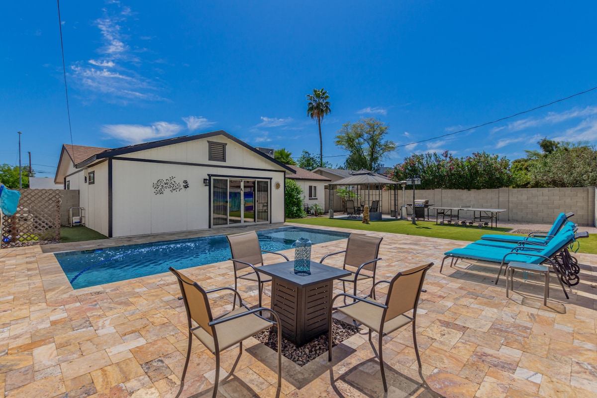An inviting outdoor space is presented, featuring a sparkling heated pool surrounded by a spacious patio laid with natural stone. Comfortable lounge chairs are positioned around the pool, and a patio table with four chairs offers a place to relax. Lush greenery and a gazebo are visible in the background.
