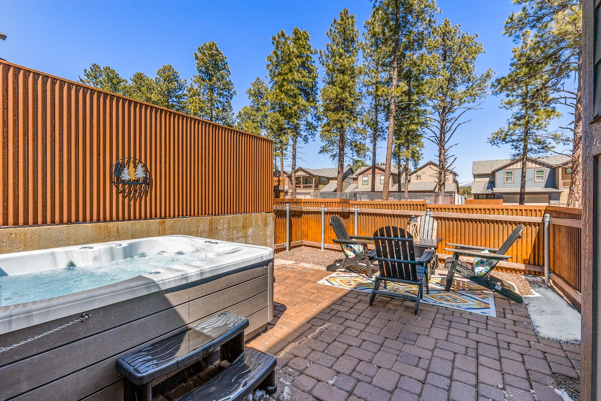 A private outdoor patio features a hot tub surrounded by wooden fencing for privacy. Comfortable seating is arranged around a fire pit on paved flooring, with tall trees visible in the background, creating a serene atmosphere for relaxation.