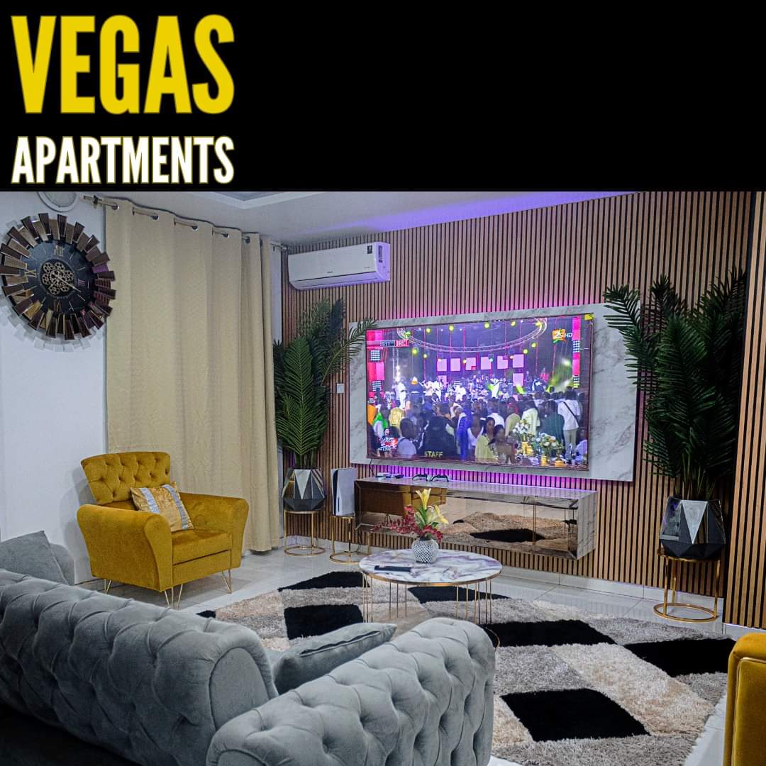 A modern living area features a plush gray sectional sofa, accent chairs in yellow, and a large flat-screen TV mounted on a textured wall. Decorative plants and stylish side tables complement the space, while a soft area rug adds warmth to the floor.