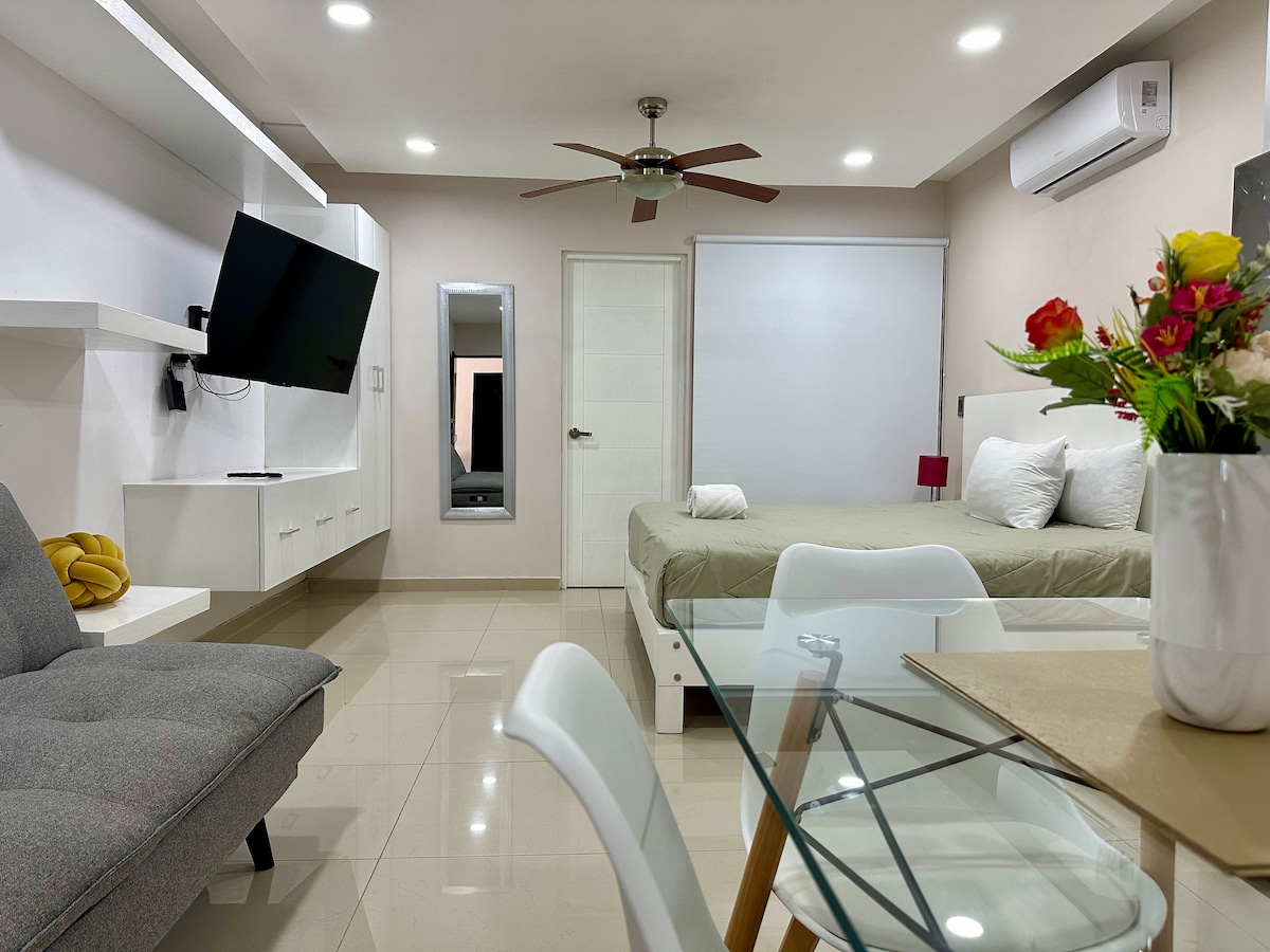 A neatly arranged studio apartment features a spacious bed and a comfortable sofa, positioned adjacent to a glass dining table. A wall-mounted TV is visible, along with a ceiling fan and ambient LED lighting. A mirror reflects the area, contributing to a bright and inviting setting.