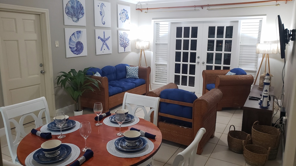 A light-filled living and dining area features a wooden dining table set for four, with blue and white dishware. Comfortable seating is provided by wicker sofas adorned with blue cushions, positioned near double glass doors leading to the outdoor patio.