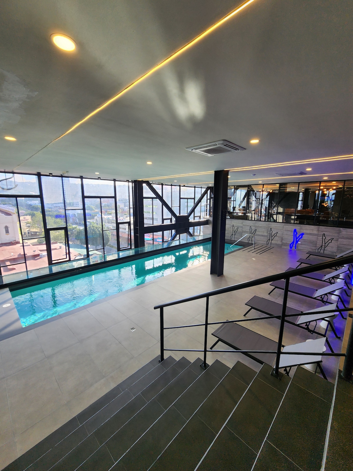 A modern indoor pool area is highlighted by large floor-to-ceiling windows, providing expansive views. The pool features a clear turquoise surface, and several sleek lounge chairs are arranged nearby for relaxation. A stairway leads down to the pool area, enhancing its spacious feel.
