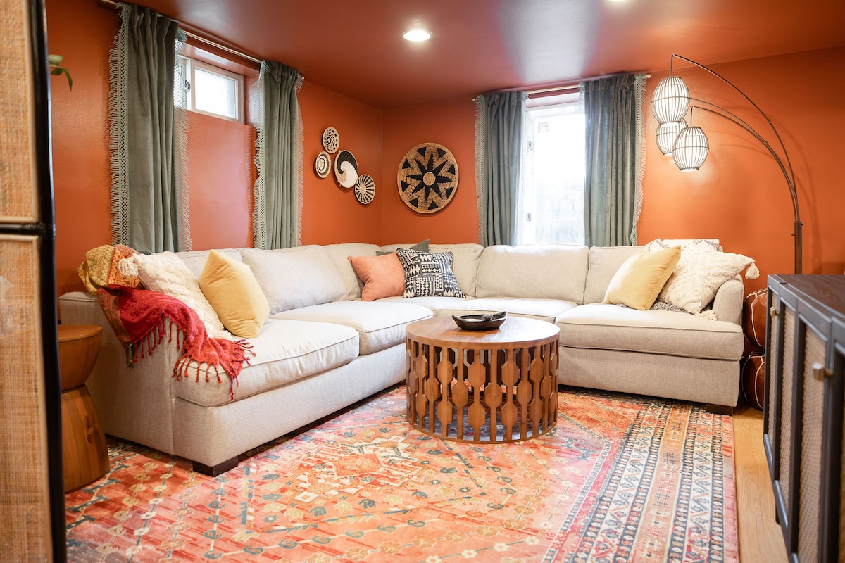 A spacious living area features a large sectional sofa adorned with decorative pillows and a soft throw. A round coffee table with a unique wood design sits in the center. Warm orange walls are complemented by light curtains, with tasteful wall decorations adding character.