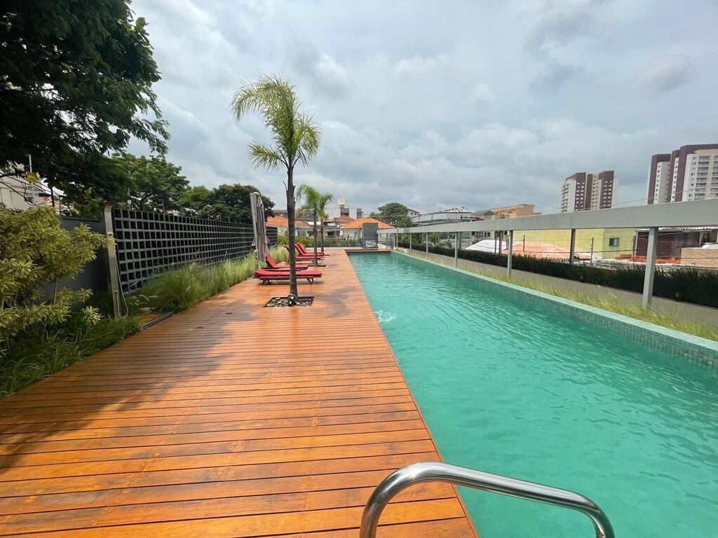 An inviting outdoor pool is surrounded by a wooden deck, featuring palm trees and lounge chairs. The pool area is well-lit by natural light, with green landscaping lining the edges, offering a serene space for relaxation.