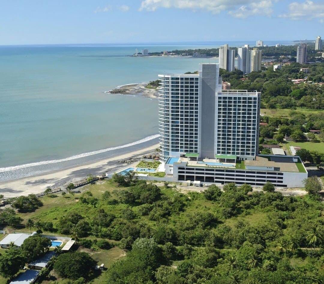 Aerial view of a modern beachfront condo building, positioned alongside a spacious coastline. The structure features multiple floors and a glass facade, offering stunning views of the ocean. Lush greenery surrounds the property, complementing the natural landscape.