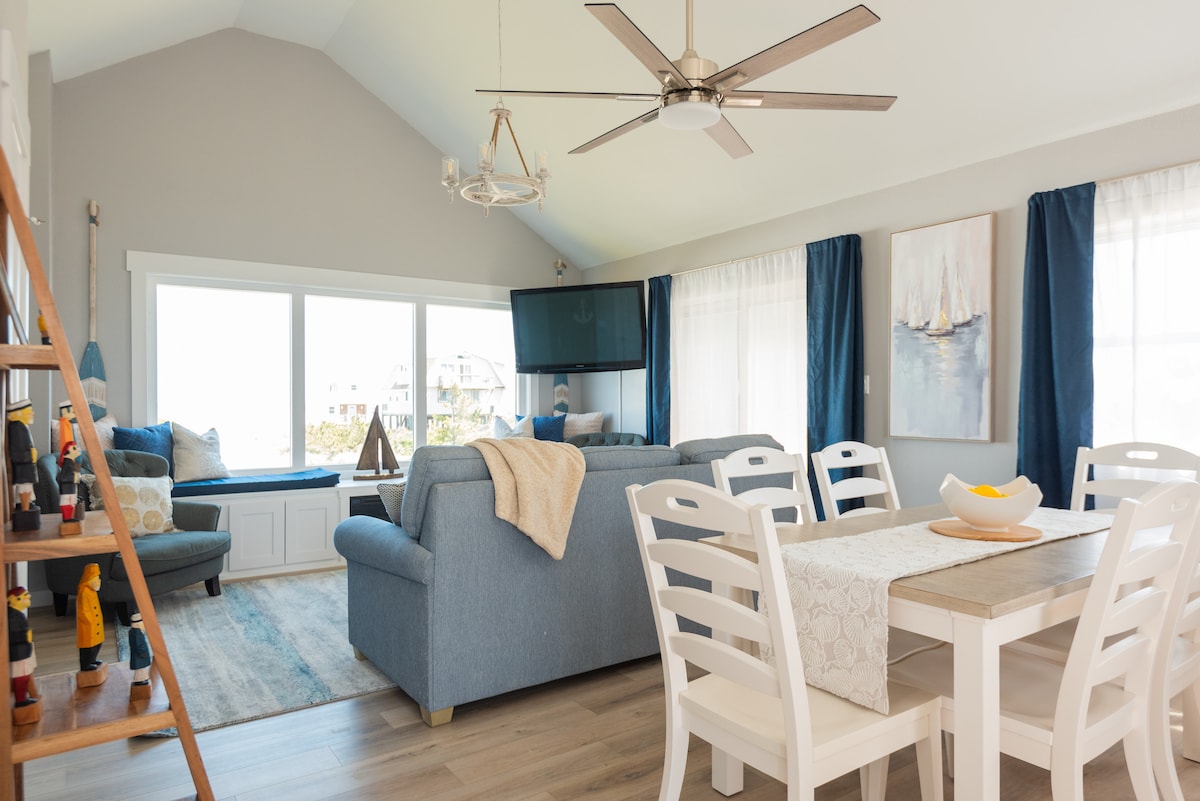 The spacious living and dining area features a light color palette with large windows allowing natural light to fill the space. A comfortable blue sofa and decorative pillows are arranged around a coffee table. A dining table with white chairs complements the inviting decor.