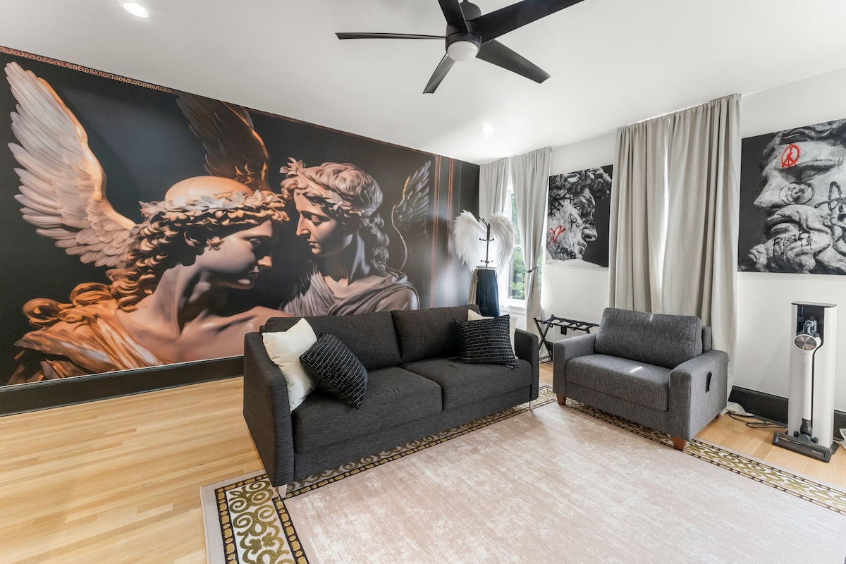 A stylish living area features a dark sofa and an armchair against a striking mural depicting an angelic embrace. Natural light filters through sheer curtains, illuminating hardwood floors and a decorative area rug that complements the modern decor.