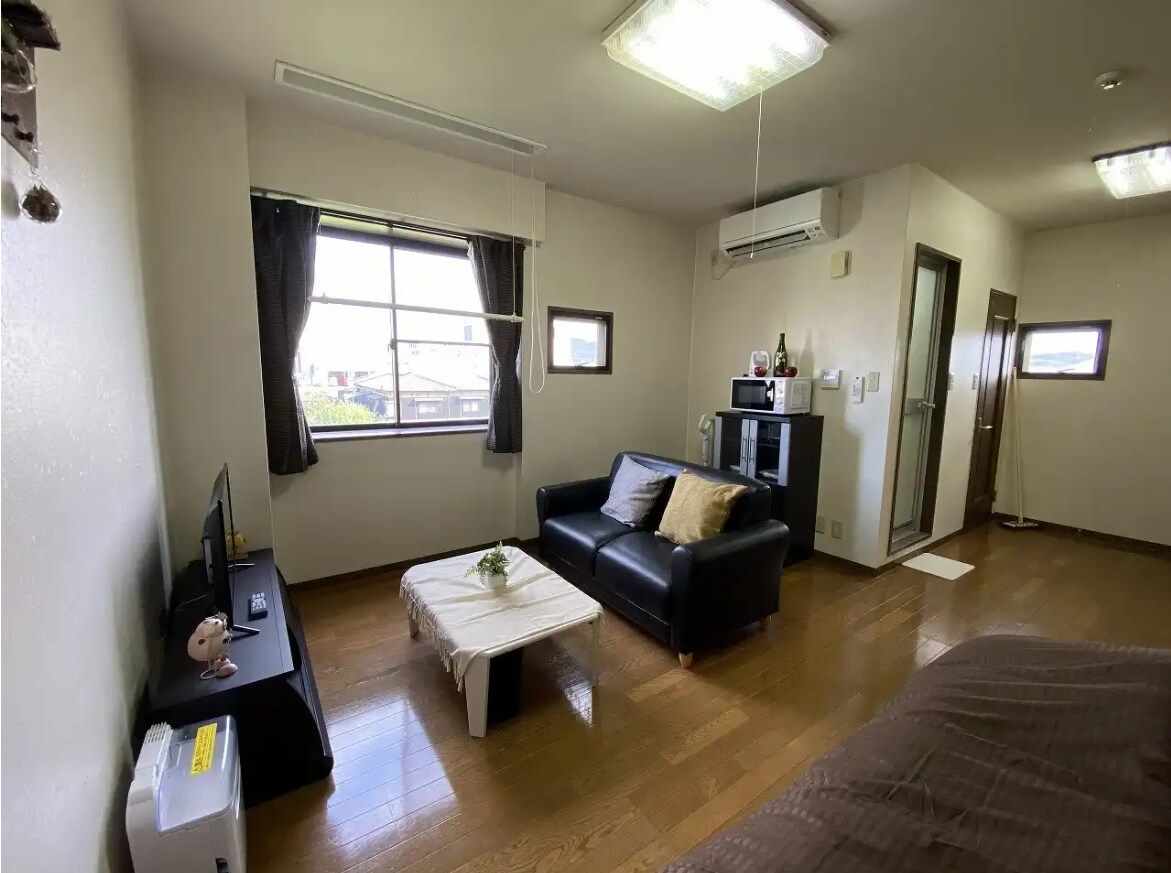 A simple living space features a black leather sofa positioned across from a low coffee table. Natural light illuminates the area through a window dressed with dark curtains. A small television sits on a stand, and air conditioning is mounted on the wall.
