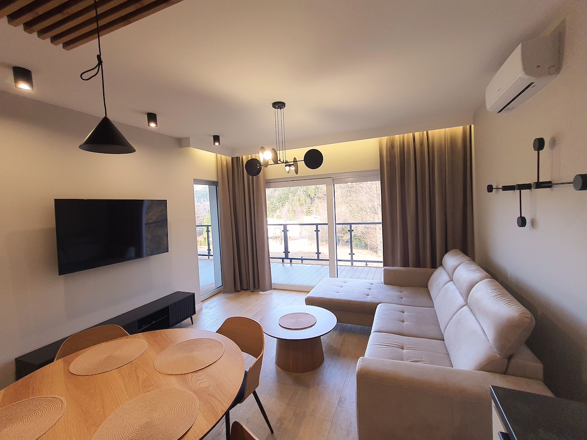 A modern living space features a light-colored sectional sofa and a round wooden coffee table. A wall-mounted television is positioned opposite the seating area. Large windows are dressed in soft curtains, allowing natural light to fill the room.