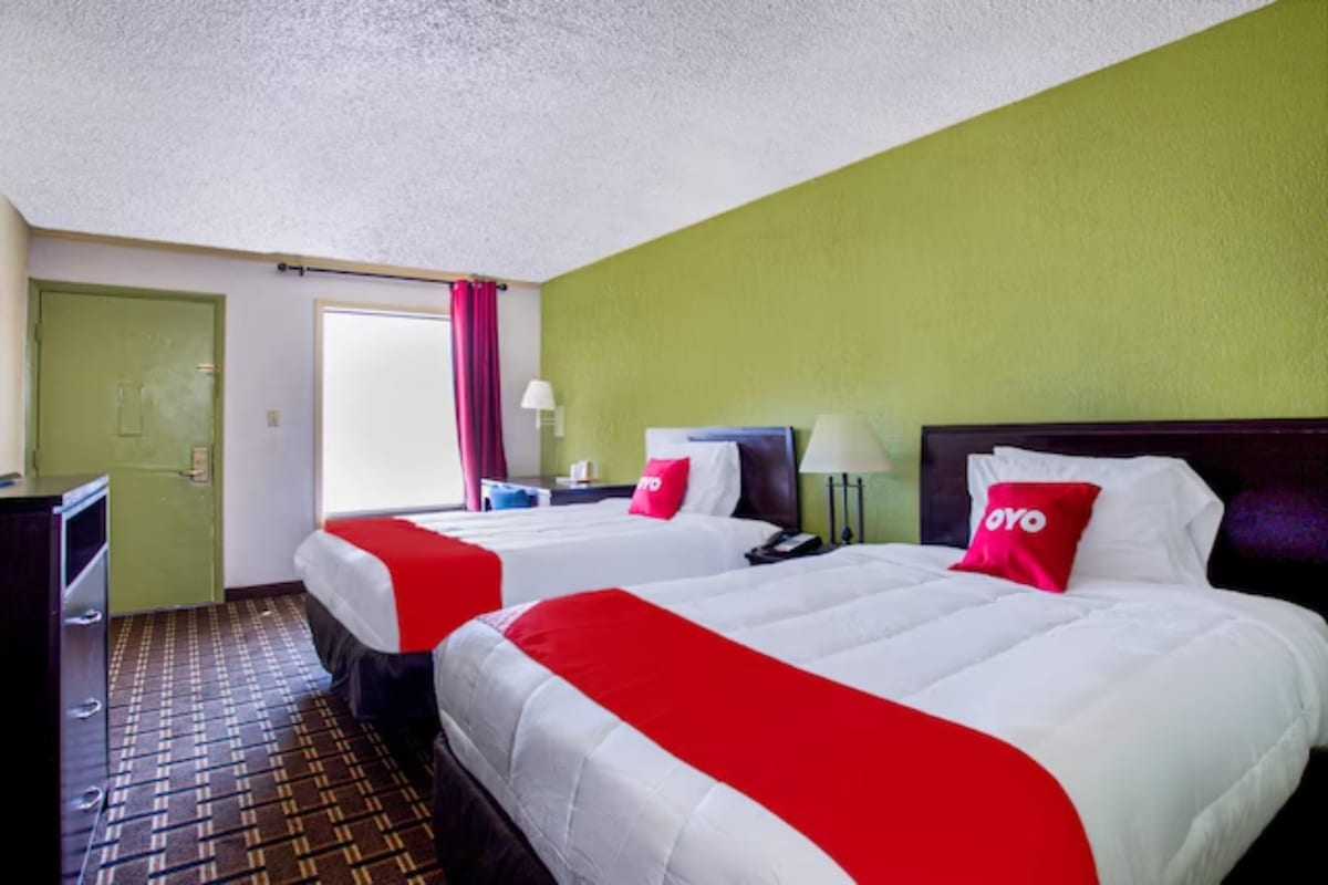 The room features two spacious beds dressed in crisp white linens, complemented by bright red accents. A dark wood dresser and a bedside lamp are present, while a window allows natural light to fill the space. A bold green wall adds a refreshing touch.