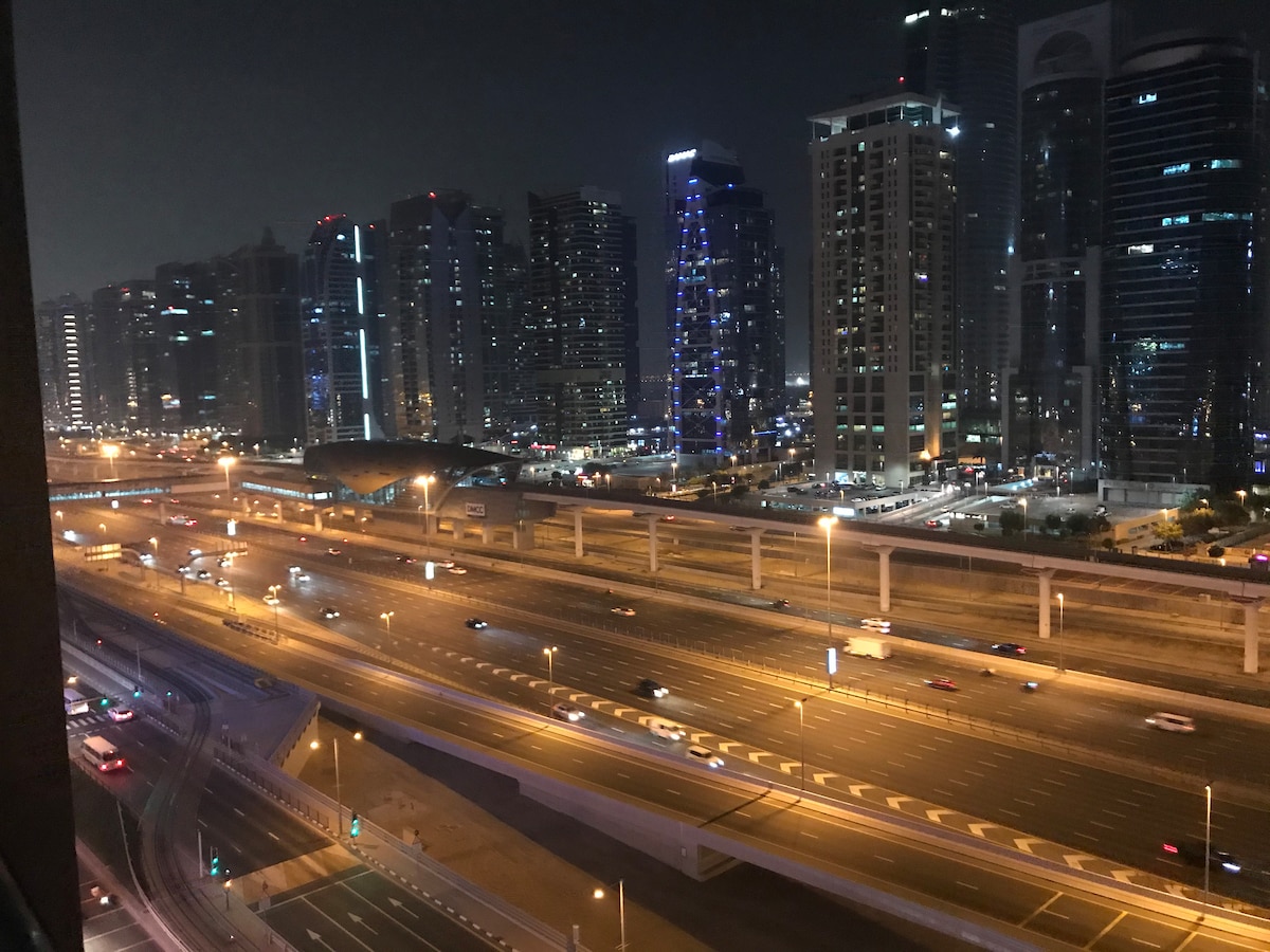 The image captures a vibrant night scene of Dubai Marina, showcasing a skyline filled with illuminated skyscrapers. Below, traffic flows on the highway, with headlights creating streaks of light. The scene reflects the energy of a bustling urban environment.
