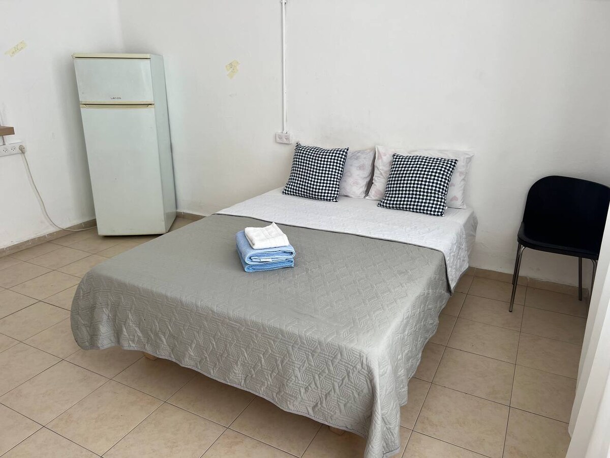 A simple sleeping area features a spacious bed covered with a light gray quilt. Two patterned pillows are positioned against the headboard. A small chair and a refrigerator are placed nearby, while neutral walls create a calm environment.