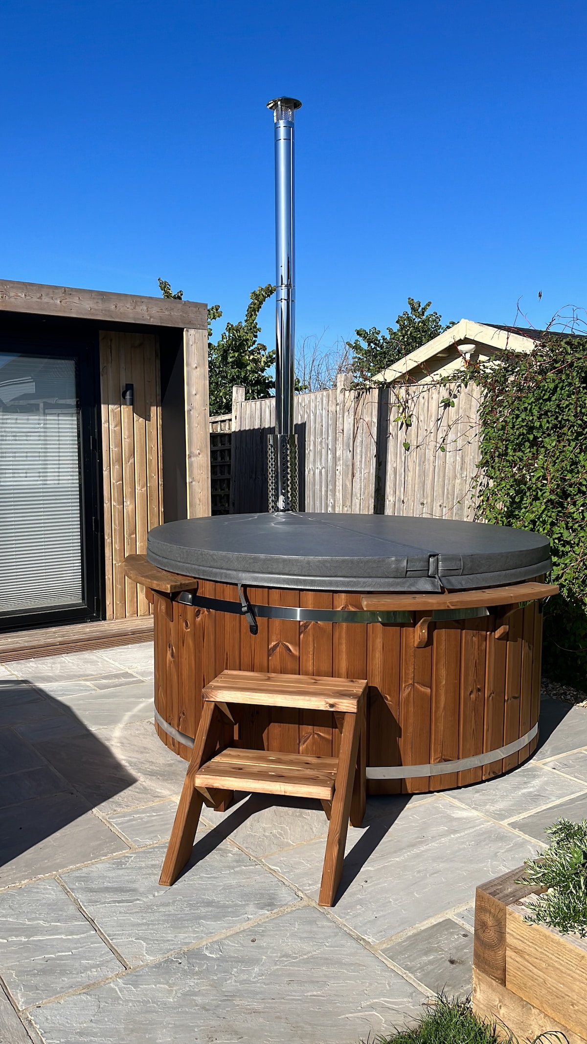 A wooden hot tub is presented on a stone patio, accompanied by a small wooden step stool for easy access. Surrounding greenery is visible, along with a wooden fence in the background, while clear blue skies define the serene atmosphere.