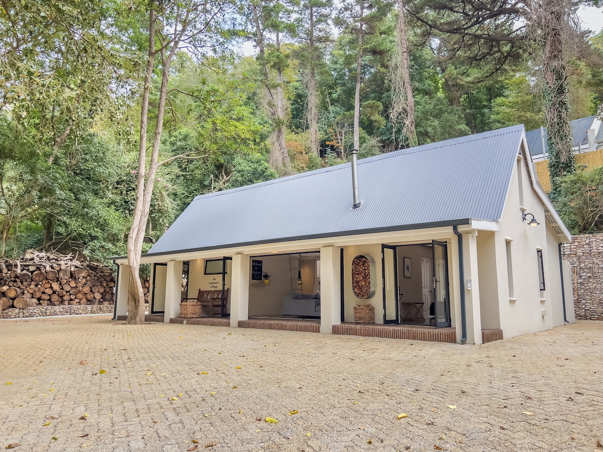 The cottage exterior showcases a modern design under a metal roof, surrounded by trees and natural landscaping. An inviting porch area with large glass doors provides access to the interior, which features a stone pathway and a backdrop of stacked firewood along the side.