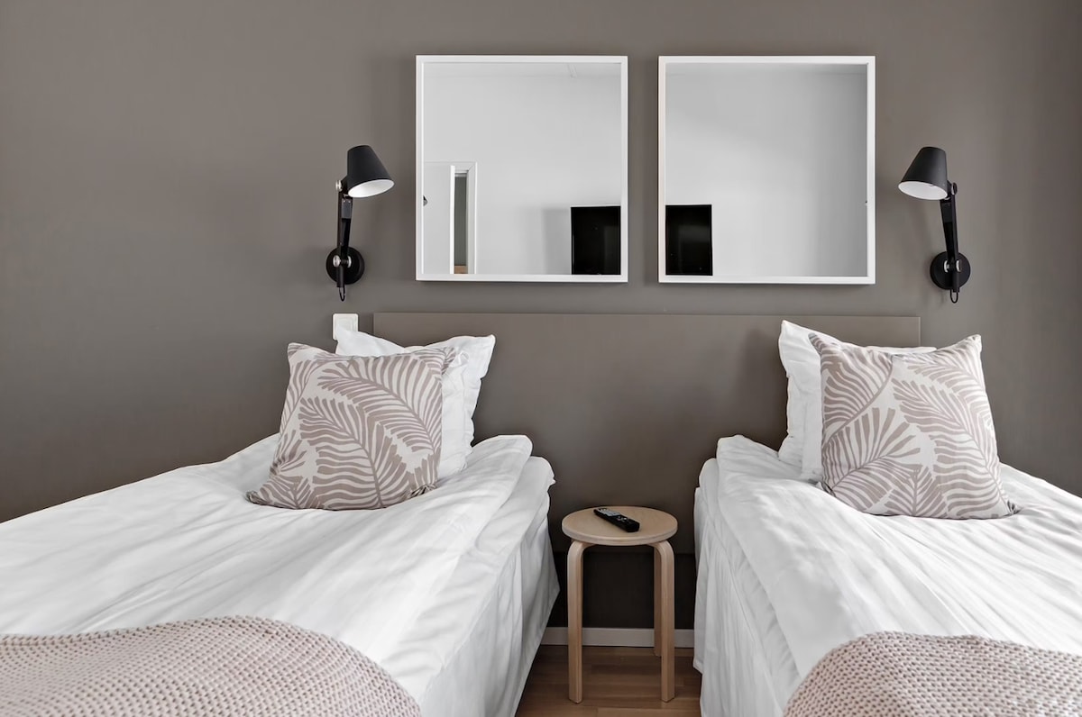 A cozy sleeping area features two single beds dressed in white linens, each with decorative pillows. Simple light fixtures are mounted on the wall, and two mirrors above the beds reflect the light. A small wooden table displays a remote control, enhancing the functional space.