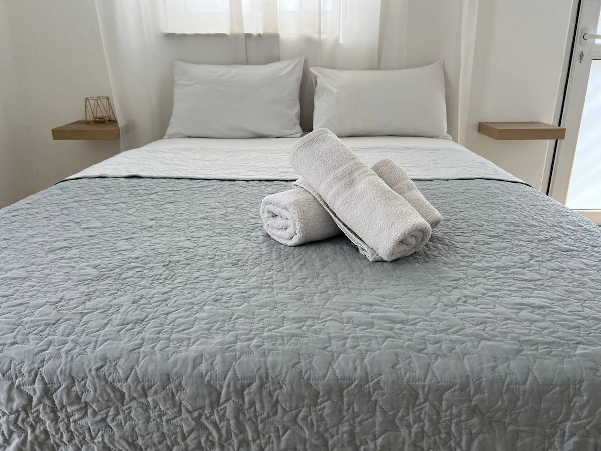 A spacious bed is dressed in a soft grey quilt with a light coverlet. White towels are neatly rolled and positioned at the foot of the bed. Natural light filters through sheer curtains, enhancing the serene atmosphere.
