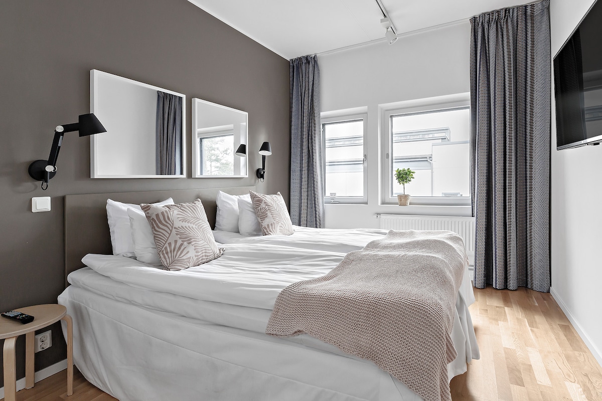 A well-lit bedroom features two single beds, which can be arranged together. Soft neutral linens are complemented by decorative pillows. Two mirrors and a TV are mounted on the walls. Light-colored curtains frame the windows, allowing natural light to brighten the space.