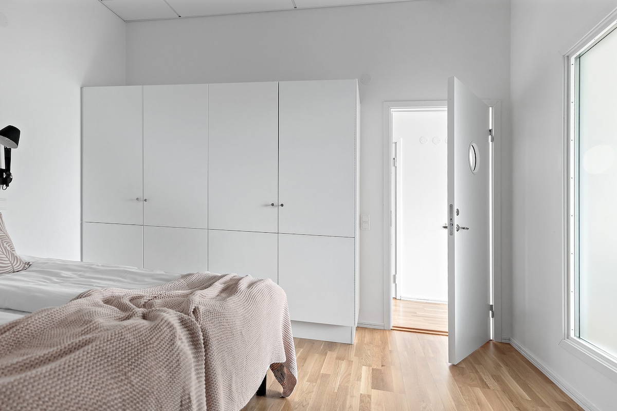 A well-lit bedroom features a spacious bed covered with a soft blanket. A sleek, white wardrobe offers ample storage, while a door opens to a small hallway. Natural light flows through a large window, highlighting the minimalist design of the room.