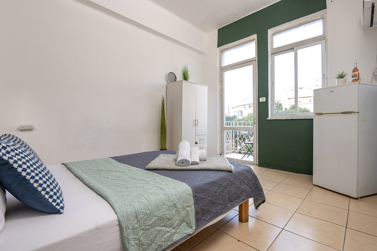 A bright and spacious room features a comfortable bed covered with a muted quilt and accented by decorative pillows. A small white wardrobe and a refrigerator are present, while natural light enters through multiple windows overlooking a balcony area.