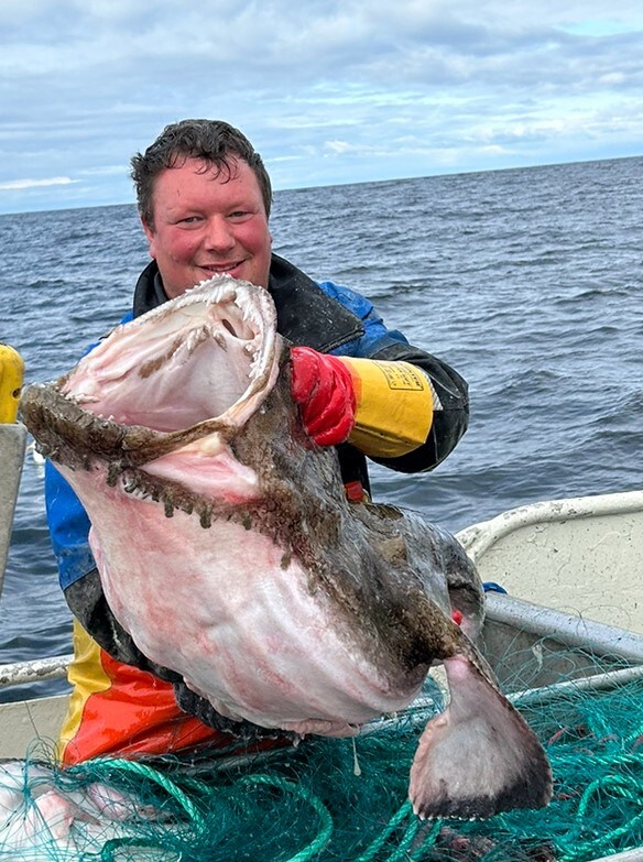 A fisherman holds a large fish, showcasing its prominent mouth and texture. The fish is displayed against a backdrop of the ocean, with gentle waves visible under a cloudy sky. Fishing nets are seen on the deck, indicating an active fishing environment.