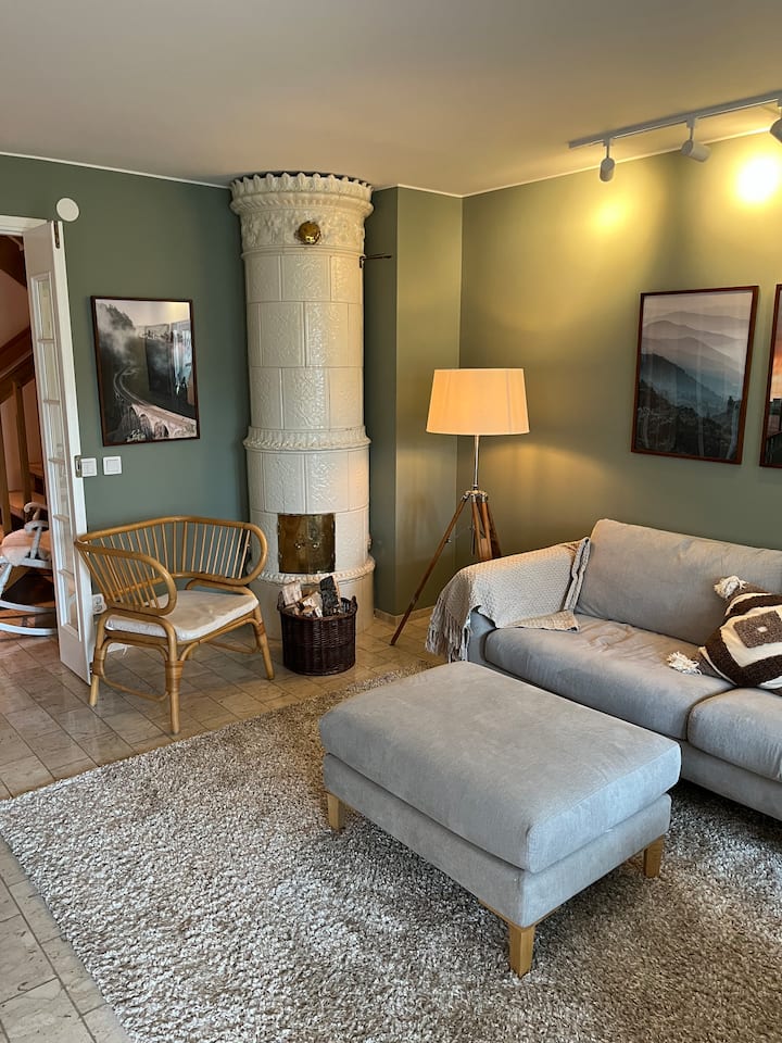 Всекидневна на приземния етаж с готварска печка с плочки, диван, фотьойли до трапезарията. 