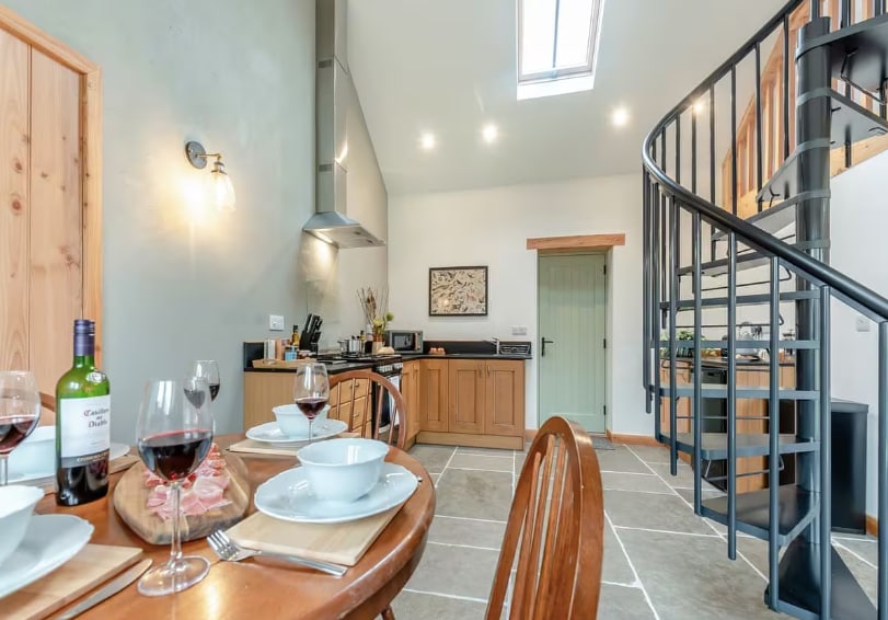 An open plan kitchen and dining area showcases exposed beams and stone flooring. A wooden dining table is set with white plates and wine glasses, complemented by a bottle of wine nearby. A spiral staircase leads to the mezzanine level, and natural light enters through a skylight.