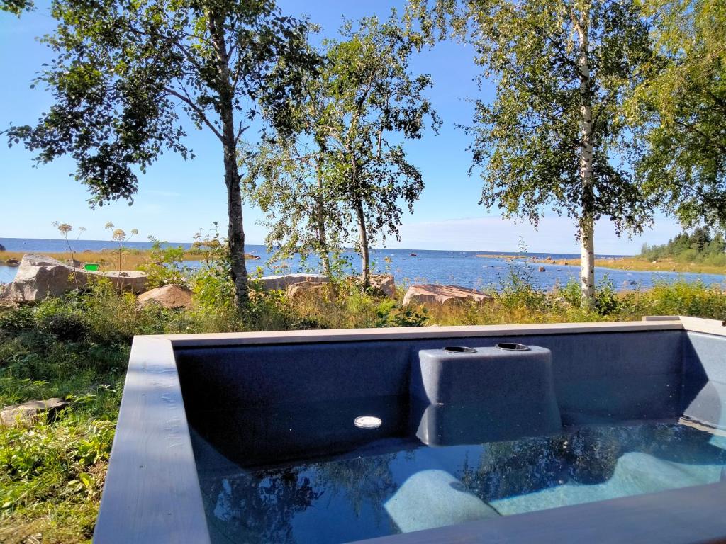 An outdoor hot tub is positioned near a serene lake, surrounded by lush greenery and trees. The water reflects the clear blue sky, with rocky shores visible in the distance. The peaceful environment highlights the natural beauty of the location.