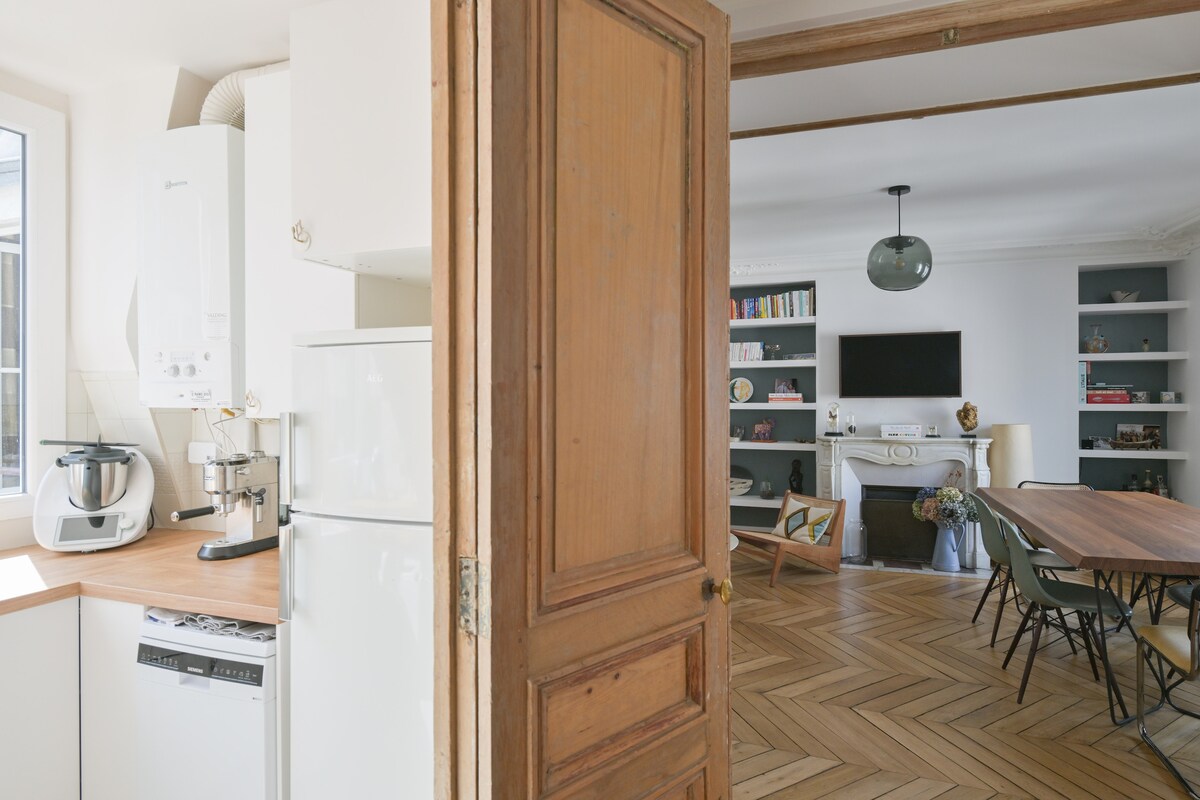 A view into the apartment showcases a well-equipped kitchen featuring modern appliances and a wooden countertop. An inviting dining area is visible beyond, with a large table surrounded by green chairs. The warm tones of the wooden door and herringbone flooring add to the overall charm.