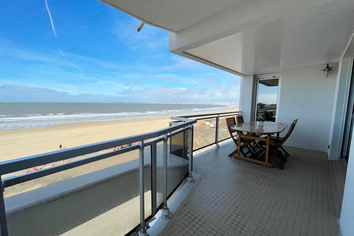 A spacious balcony extends from the apartment, offering an unobstructed view of the ocean and beach. A wooden table and chairs are arranged for dining, with railings ensuring safety. The sky is bright with clouds, and the waves can be seen gently rolling ashore.