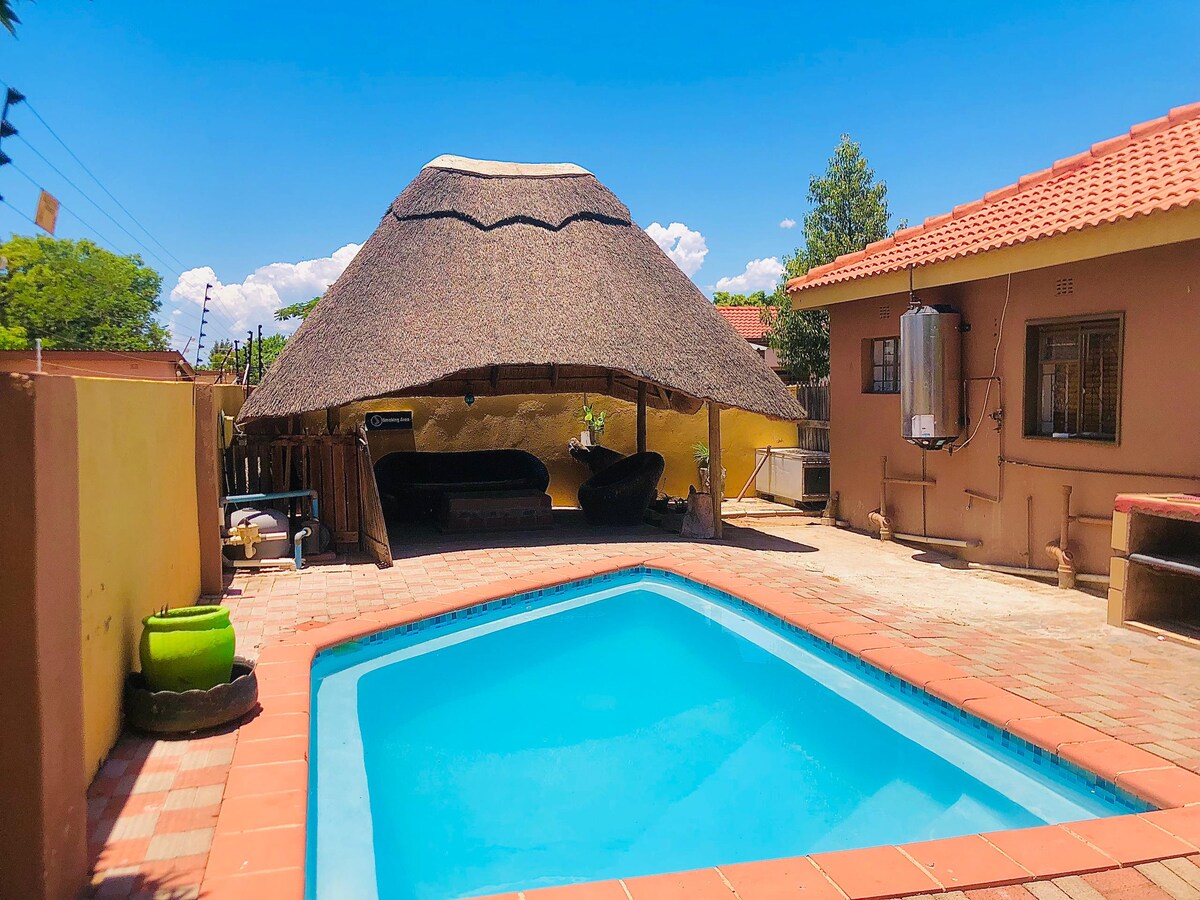 A refreshing pool is framed by a tiled patio, with a thatched-roof gazebo providing shaded seating nearby. The surrounding area is marked by sunny blue skies and green trees, while a neighboring building is visible, contributing to the outdoor relaxation space.